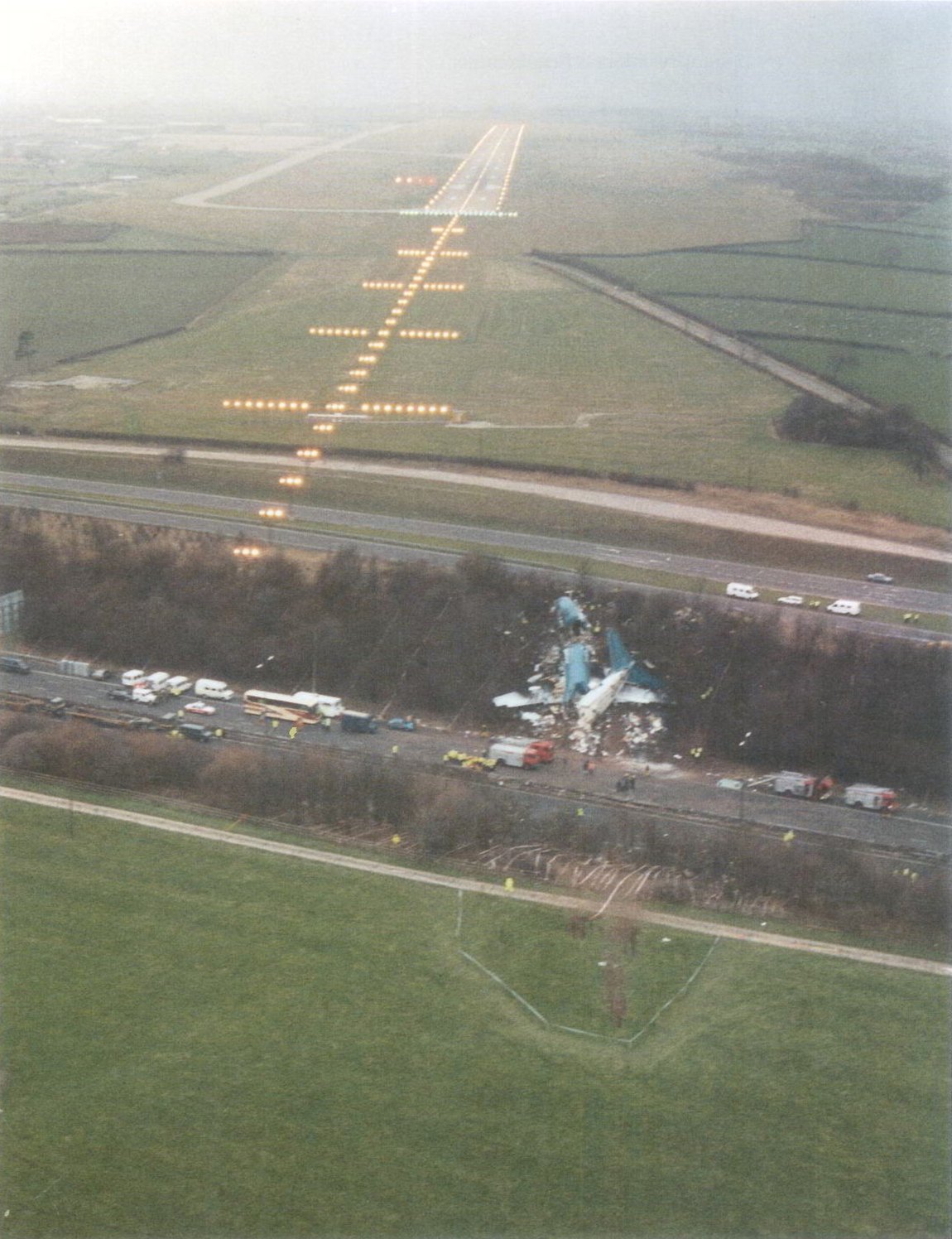Aerial photograph taken by the Air Accidents Investigation Branch showing the broken British Midland 737 on the M1 embankment, with the motorway, the approach lighting and the lit runway of East Midlands Airport in line beyond it