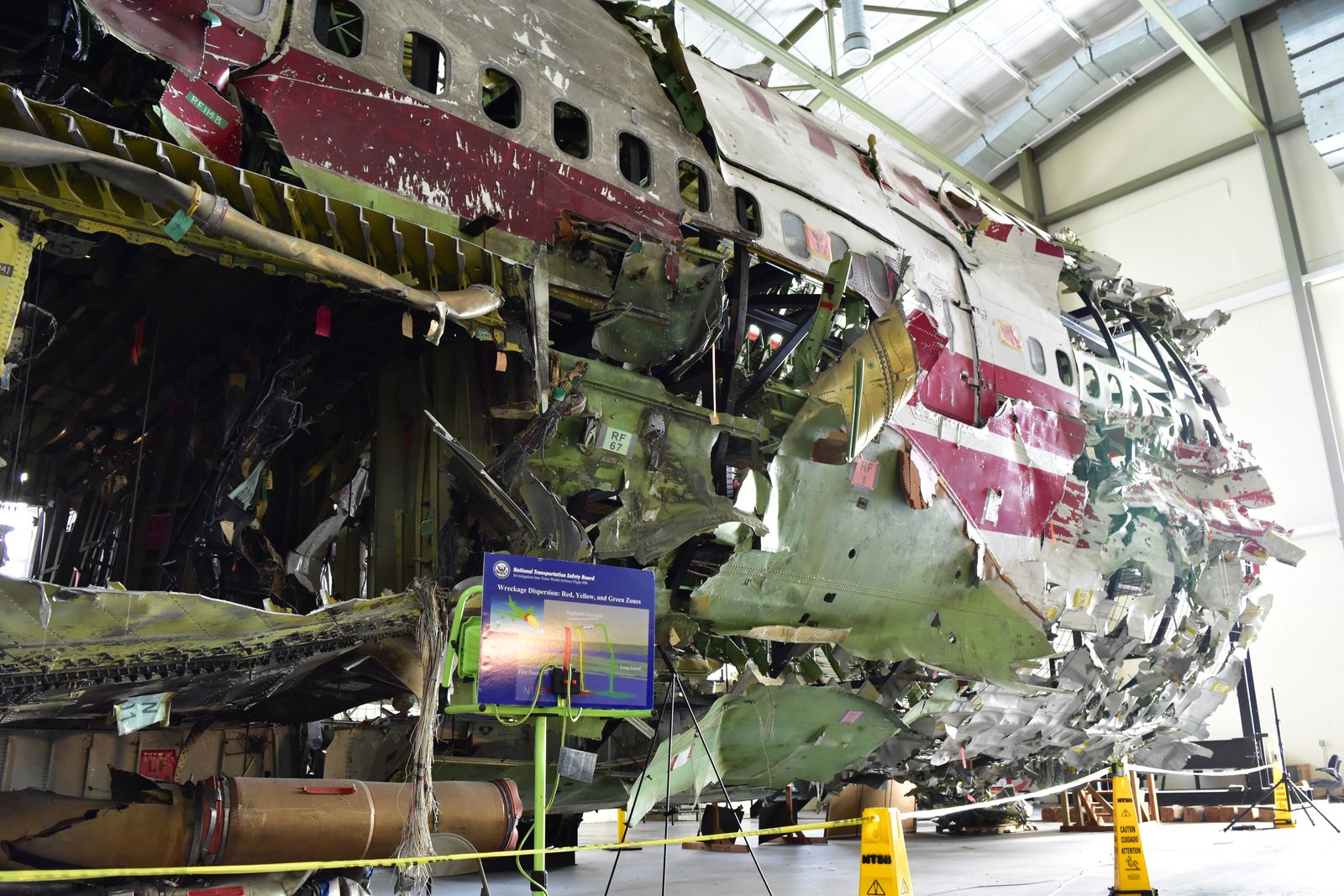 The reassembled forward fuselage of TWA Flight 800 in the NTSB training centre hangar, showing numbered wreckage pieces on a steel frame and an NTSB display board about wreckage dispersion zones